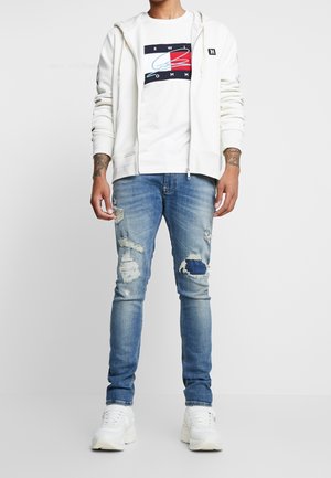 Sudadera blanca con cremallera sobre una camiseta gráfica blanca, combinada con unos vaqueros azules desgastados. Con zapatillas blancas. Outfit casual.