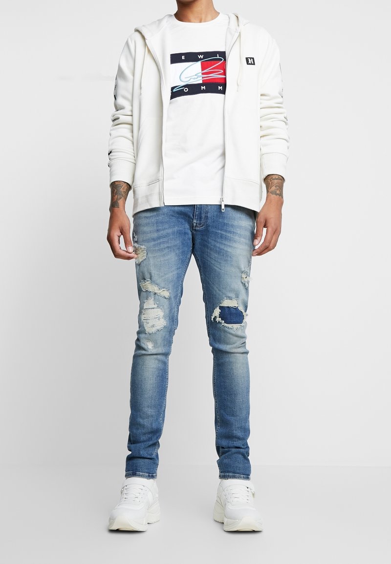 Sudadera blanca con cremallera sobre una camiseta gráfica blanca, combinada con unos vaqueros azules desgastados. Con zapatillas blancas. Outfit casual.