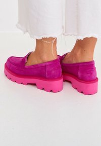 En person som bär vita byxor med fransat fåll och klarröda mockaloafers med tjocka sula, stående på ett vitt golv.