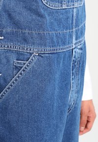 Keskisinis denim overall, millel on eesmine tasku, õmblusteadus ja lõdvem sobivus. Kangas on sile, veidi tekstureeritud viimistlusega.