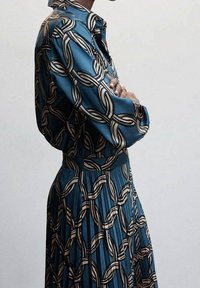 Robe turquoise avec une jupe plissée, présentant un motif audacieux en chaîne alternée beige et noire. Tissu lisse avec un col chemise.