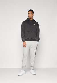 Sweat-shirt gris avec capuche et poche avant, arborant un logo blanc. Pantalons de survêtement gris clair avec texte et poignets bleu marine. Des baskets blanches complètent le look.