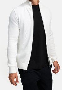 흰색 니트 지퍼 재킷, 질감이 있는 패턴, 리브드 커프스와 밑단이 있으며, 검은색 티셔츠 위에 걸쳤습니다. 디자인 및 소재의 자세한 부분이 강조되었습니다.