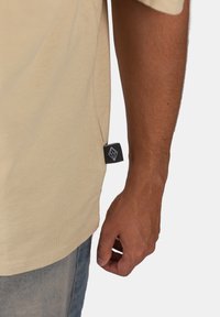 Béžové bavlněné tričko s krátkým rukávem, s božní etiketou v černé barvě a geometrickým bílým logem. Viditelné švy a měkká textura.