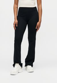 Femme portant un pantalon noir ajusté et des baskets blanches épaisses, debout devant un fond uni, les mains détendues le long du corps.