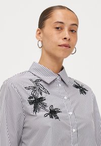 Striped button-up shirt with black floral beaded embroidery. Features a pointed collar and white buttons, made from a lightweight fabric.