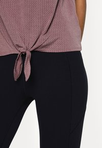 Femme portant un haut mauve texturé noué à la taille avec un legging noir moulant, debout devant un fond blanc uni.