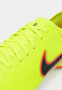 Neón žlté futbalové topánky s textúrovaným povrchom, čiernym a oranžovým Nike Swoosh, plochými šnúrkami a aerodynamickým dizajnom. Ľahký a syntetický materiál.