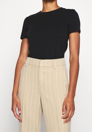 Femme portant un haut noir ajusté à manches courtes, rentré dans un pantalon beige taille haute à rayures verticales blanches.
