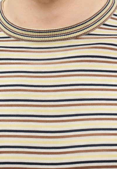 Κοντινό πλάνο ενός στρογγυλού λαιμοδέτη με ριμπ ύφασμα και ριγέ πολύχρωμο πλεκτό σχέδιο σε αποχρώσεις μπεζ, καφέ, μαύρο και κίτρινο.