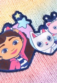 Un patch en tissu coloré représentant une fille aux cheveux bouclés et un personnage de chat, entourés d'étoiles, cousu sur un fond en tricot pastel.