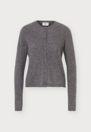 Le mannequin se tient devant un fond clair et uni, vêtu d’un cardigan gris boutonné sur le devant, d’un pantalon noir à jambes larges et de chaussures noires.
