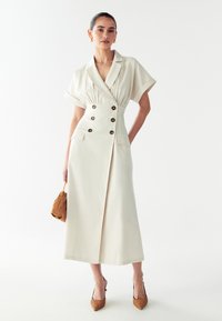 Abito in lino beige con maniche corte e larghe, doppio petto, due tasche laterali e una silhouette dritta, abbinato ad accessori marroni.