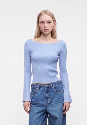 Ung kvinde med langt blond hår iført en lyseblå ribbet off-shoulder sweater og højtaljede blå jeans står foran en hvid baggrund.