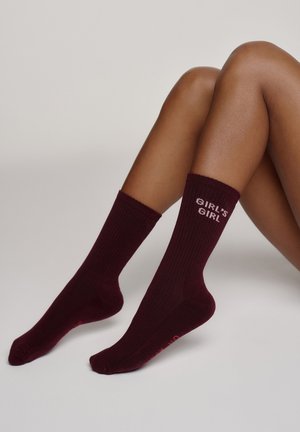Nogi w ciemnofioletowych prążkowanych skarpetkach z napisem "GIRL'S GIRL" u góry na jasnoszarym tle.