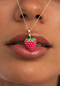 Nahaufnahme von Lippen, die eine goldene Halskette mit einem rot-grünen, erdbeerförmigen Anhänger halten.