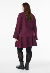 Kobieta z długimi brązowymi włosami, ubrana w długą purpurową sukienkę z długim rękawem, czarne rajstopy i czarne botki za kostkę, stojąca tyłem na białym tle.