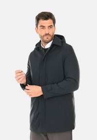 Cappotto nero con cappuccio realizzato in tessuto liscio, presenta una chiusura frontale con bottoni, tasche laterali e polsini regolabili. Il design è semplice e lineare.