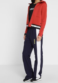 Mujer vistiendo una sudadera roja con cremallera, una camiseta negra de Calvin Klein y pantalones deportivos azul marino con rayas blancas en los laterales y zapatillas negras.