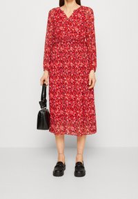 Robe rouge à fleurs avec manches longues, col en V, taille smockée et longueur mi-mollet. Sac à main noir texturé et chaussures noires épaisses complètent la tenue.