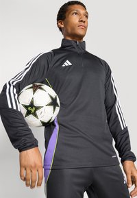 Black athletic top with purple side accents, white Adidas logo, long sleeves, and a zip collar, held with a patterned soccer ball.