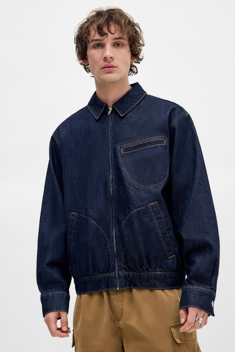 Donkerblauwe denimjack met een ritssluiting aan de voorkant, een kinband en een elastische onderrand. Voorzien van een patch zak en gebogen zijzakken met gele stiksels.