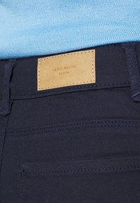 Jeans de mezclilla azul oscuro con una etiqueta de cuero marrón claro con la inscripción "VERO MODA" y "DENIM" en relieve. Presenta bolsillos cosidos y una textura suave.