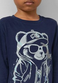 Ein Kind trägt einen marineblauen Sweatshirt mit einem glänzenden silbernen Grafik eines Bären in Sonnenbrille und einer Kappe mit Kapuzen-Design.
