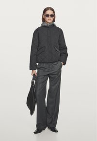 Black quilted jacket with a gray fur-lined hood, paired with gray wide-leg trousers. Model holds a black handbag.