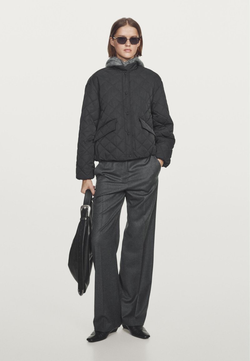 Black quilted jacket with a gray fur-lined hood, paired with gray wide-leg trousers. Model holds a black handbag.