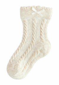 Calza creata a maglia di colore crema con motivi a treccia, bordo a costine e un piccolo fiocco bianco come dettaglio. Realizzata in morbido filato testurizzato.