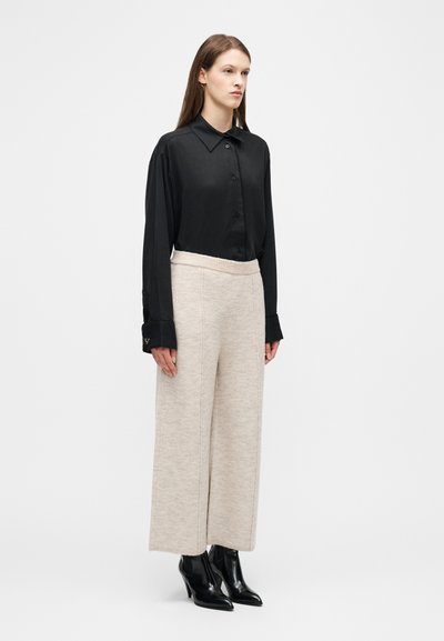 Γυναίκα που φοράει μια φαρδιά μαύρη πουκάμισα με κουμπιά, μπεζ πλεκτό παντελόνι φαρδιάς γραμμής και μαύρα μυτερά μπότες αστραγάλου με τακούνι, στέκεται μπροστά σε μια απλή φόντο.