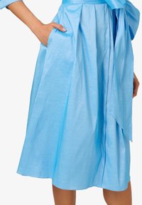 Abito azzurro chiaro, lungo fino al ginocchio, con una texture setosa, design plissettato e tasche laterali. Presenta una vita legata e una silhouette fluida.