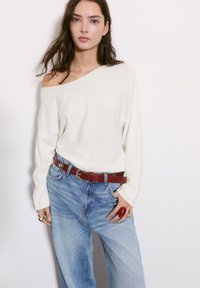 Biela off-shoulder top s dlhými rukávmi v kombinácii s vysokými modrými širokými džínami. Červený kožený opasok a prsteň dodávajú odvážne akcenty.