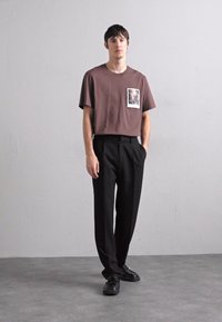 Barna, rövid ujjú póló elülső zsebnyomattal, fekete nadrággal és fekete cipővel párosítva. Egyszerű dizájn, laza fit.