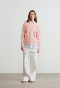 Γυναίκα που φοράει ανοιχτό ροζ φούτερ πάνω από μπλε ριγέ πουκάμισο και άσπρο παντελόνι φαρδύ, στέκεται μπροστά σε λευκό τοίχο.