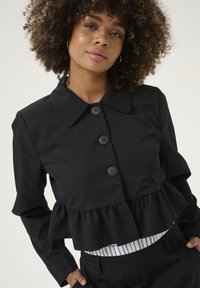 Musta blusa, jossa on peplum-muotoilu, pyöreä kaulus ja neljä nappia. Kangas on sileä ja muoto on jämäkkä. Raidallinen aluspaita näkyy.
