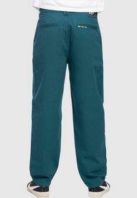 Pantaloni in cotone turchese con design a gamba dritta, due tasche posteriori e un accento arcobaleno sulla tasca destra; abbinati a sneakers in bianco e nero.