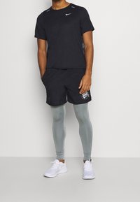 T-shirt noir à manches courtes Nike avec accents réfléchissants, associé à un short noir avec un motif. Leggings gris clair et baskets blanches.