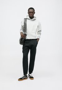 Szary dres z kapturem, czarne spodnie dresowe i czarne sneakersy z białymi sznurowadłami oraz gumową podeszwą. Model trzyma czarną torbę z teksturowanym paskiem.