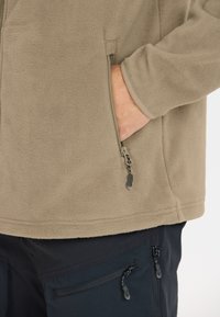 Beige fleece-takki, jossa on teksturoitu pinta, varustettuna vetoketjullisella taskulla, sekä kontrastiväri mustat housut vetoketjullisilla taskuilla.