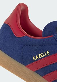 Blå ruskindsneakers med røde læderdetaljer, røde striber, gummisål og "GAZELLE" i gylden skrift på siden.