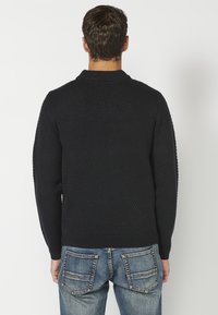 Dunkelblauer Strickpullover mit strukturiertem Muster, Rundhalsausschnitt und gerippten Bündchen. Kombiniert mit blauen Jeans mit hinteren Taschen.