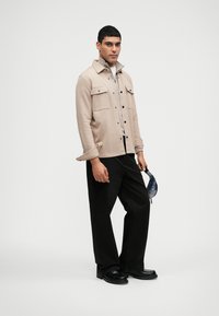 Giacca camicia beige con bottoni neri, due tasche frontali e colletto pieghevole, abbinata a pantaloni neri a gamba larga e scarpe nere.