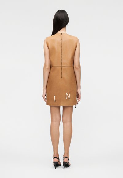 Γυναίκα με μακριά σκούρα μαλλιά που φοράει αμάνικο ταμπά δερμάτινο μίνι φόρεμα με φερμουάρ στο πίσω μέρος και μαύρα σανδάλια με ψηλό τακούνι, γυρισμένη από την πίσω πλευρά.