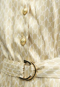 Μπεζ ύφασμα με επαναλαμβανόμενο λευκό σχέδιο λογοτύπου, με χρυσά κουμπιά και αγκράφα ζώνης. Λεία υφή σατέν, ραμμένο σχέδιο.