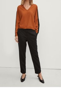 Suéter naranja de cuello en V con mangas negras, combinado con pantalones negros ajustados y zapatos negros de punta, exhibido sobre un fondo neutro.