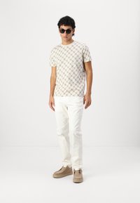 Chemise à manches courtes beige avec un motif géométrique, assortie à un pantalon blanc et des chaussures marron clair. Accessoirisée avec des lunettes de soleil noires.