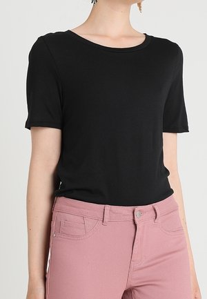 Kvinde iført en ensfarvet sort kortærmet t-shirt, der er stoppet ned i højtsiddende lyserøde bukser med forlommer og bæltestropper.