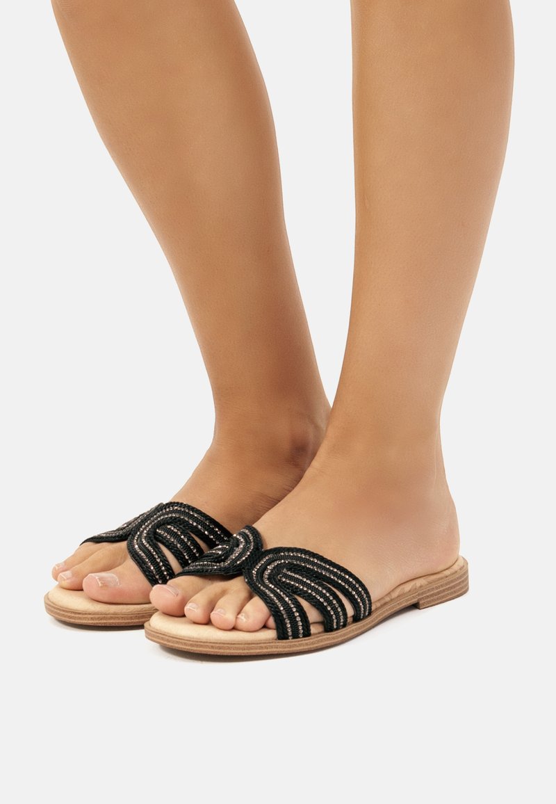 Des sandales noires à semelle plate avec un design tressé, présentant des accents métalliques et mettant en avant une texture semblable à du cuir lisse.